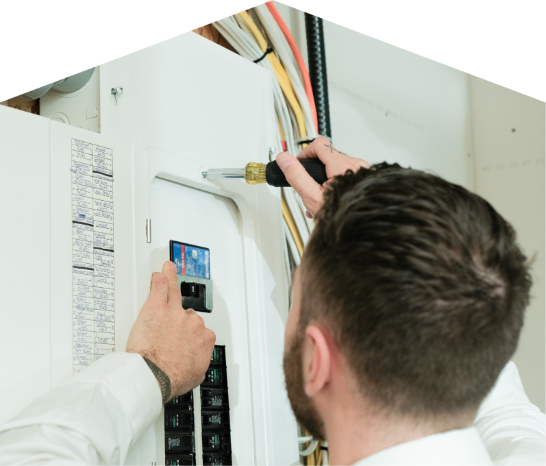 Peel electrician using a screwdriver to perform an electrical panel upgrade, working near organized wiring in a residential wall.