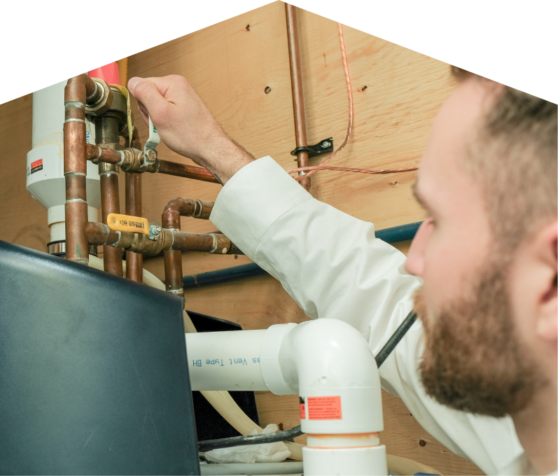 Peel water heater specialist tightens a valve on copper piping during a water heater installation, with exposed wood paneling and wiring in the background. Peel water heater specialist tightens a valve on copper piping during a water heater installation, with exposed wood paneling and wiring in the background.